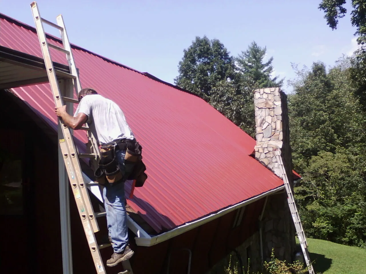 Roofer on a ladder beside a red metal roof and stone chimney — Soffit & Fascia Repair by Roofing Brothers in Bushkill